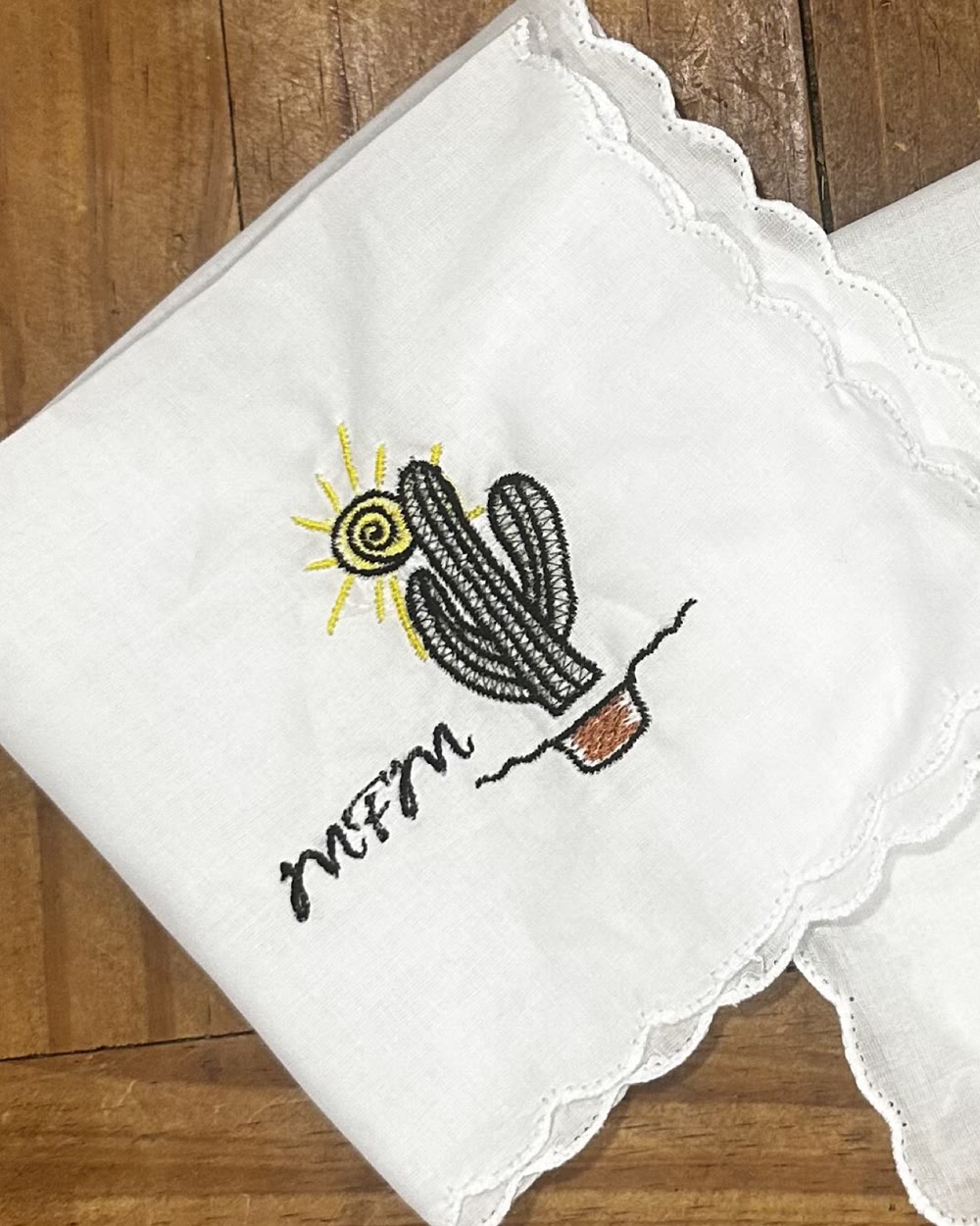 A white scalloped-edge handkerchief embroidered with a potted cactus, a yellow sun and a script monogram