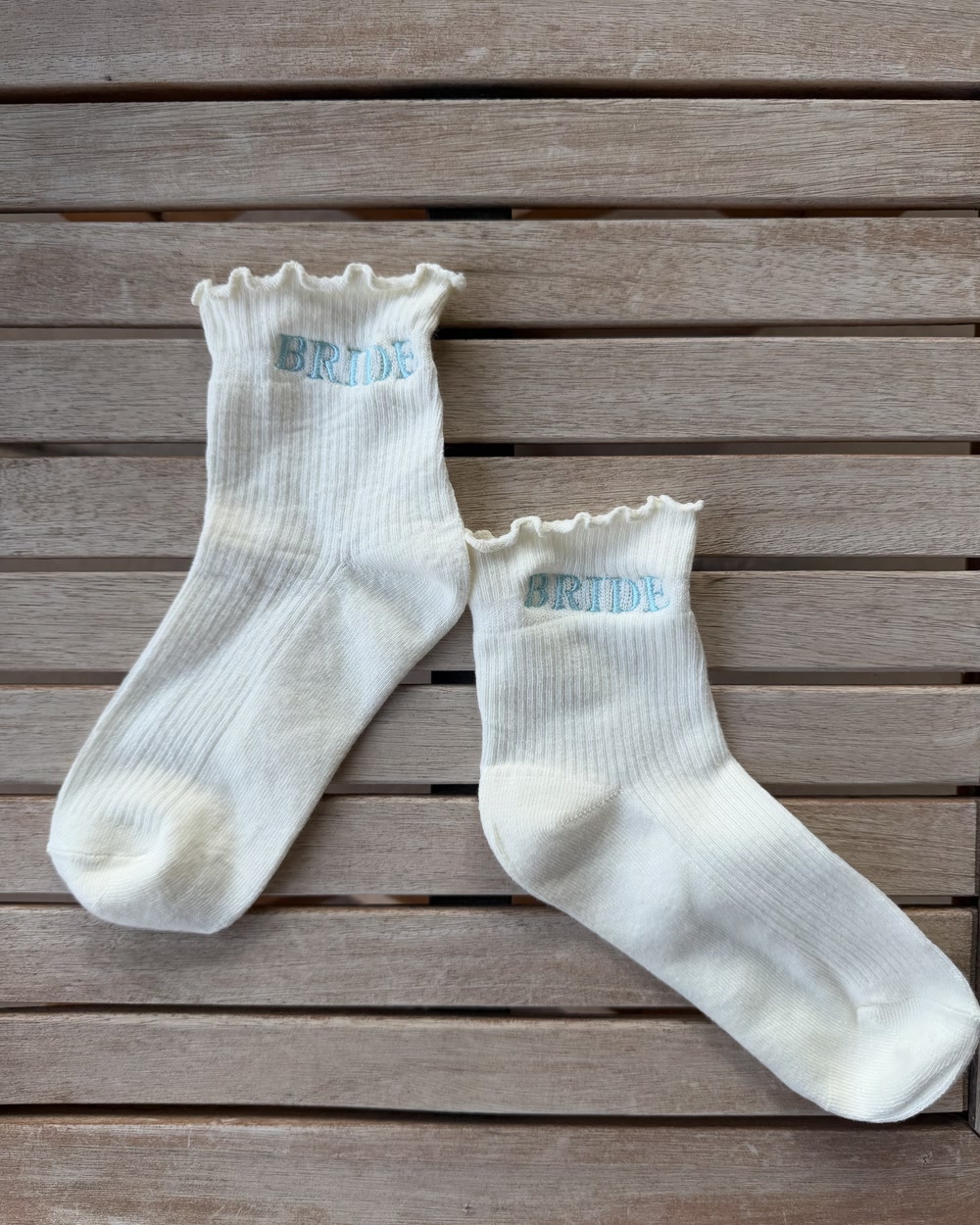 A pair of ivory ruffle socks embroidered with the word BRIDE in pale blue